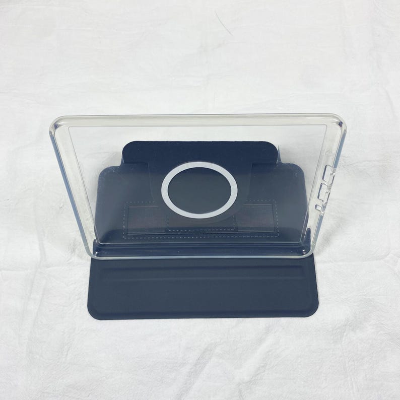 Puede incluir: Una funda protectora transparente para tableta con una base de soporte plegable en color azul oscuro. La parte posterior incluye un anillo de carga magn&eacute;tico circular centrado en un panel azul oscuro, con recortes para botones laterales.