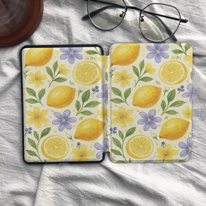 May include: A protective e-reader case featuring a repeating pattern of yellow lemons, lemon slices, small yellow flowers, light purple flowers, and green leaves on a cream background. The case is open, displaying the interior and exterior design.