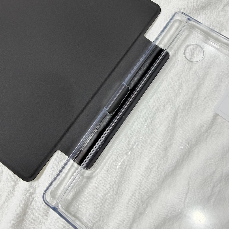 Puede incluir: Una funda protectora para tableta que presenta una cubierta negra mate unida a una carcasa trasera transparente. El dise&ntilde;o incluye una secci&oacute;n de bisagra negra dedicada para el soporte del dispositivo, con una construcci&oacute;n funcional.