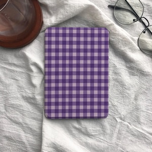 May include: A rectangular coaster or notebook featuring a purple and light purple gingham pattern. The item is placed on a white, textured fabric. A glass and glasses are partially visible in the background.