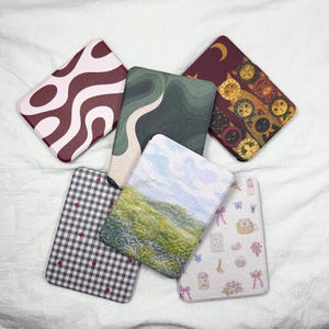 Op de afbeelding: Een verzameling rechthoekige hoesjes met verschillende ontwerpen. Eén hoesje heeft een golvend patroon in bordeauxrood en roze. Een andere heeft een abstract ontwerp in groen en beige. Andere hoesjes tonen een landschap, een ruitpatroon en een kattenontwerp.