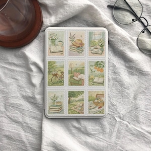 May include: A white rectangular tin featuring nine postage stamp-style illustrations. Each stamp depicts a cozy scene with books, flowers, and outdoor settings, including a bicycle and picnic basket. The illustrations are in soft pastel colors.