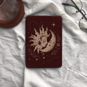 May include: A rectangular dark maroon e-reader cover featuring a cream-colored line art illustration of a sun and crescent moon with human faces, surrounded by delicate botanical vines, small stars, and constellations on the front surface.
