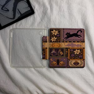 May include: A clear plastic e-reader case featuring a rectangular cover with a printed design of a black cat, a sun, clouds, potion bottles, flowers, a moth, and a heart. The design uses a palette of purple, orange, and cream colors.