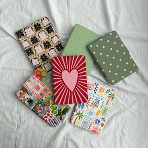 May include: A collection of colorful, patterned notebooks. Designs include floral, polka dot, heart, and travel-themed covers. The notebooks are arranged on a white, textured surface.