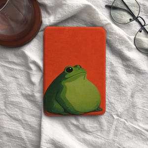 Puede incluir: Una funda rectangular para lector electrónico con un fondo naranja vibrante y una ilustración centrada de una rana verde y regordeta con grandes ojos negros. La rana aparece en posición sentada, ocupando la parte inferior.
