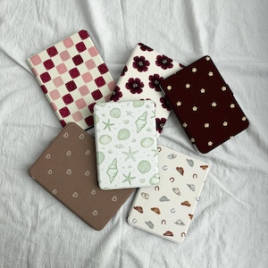Może przedstawiać: Kolekcja prostokątnych etui z różnymi wzorami. Wzory obejmują różowo-bordową kratę, bordowe kwiaty, brąz z małymi białymi kwiatami, brąz z sercami i wzór muszli.