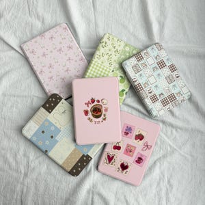 Puede incluir: Cinco fundas rectangulares para lectores electrónicos con varios diseños: una funda rosa claro con lazos y patos blancos, una funda verde con flores y cuadros, una funda a cuadros azules y marrones con caras de gato, una rosa con fresas y pegatinas.