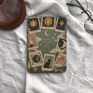 Peut inclure: Une housse de liseuse rectangulaire avec un fond vert orné d'illustrations de cartes de tarot, dont Le Soleil, La Force, L'Étoile, Le Magicien, La Lune, Le Monde et Le Cœur. Le texte indique, Believe in the magic of the season.