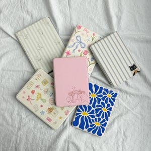 May include: A collection of decorative tablet cases in various designs. Cases feature floral, striped, and illustrated patterns, including a pink case with a champagne toast design and a blue daisy pattern.