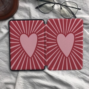Puede incluir: Una funda rectangular para lector electr&oacute;nico con fondo rojo oscuro y un coraz&oacute;n rosa claro centrado en cada lado. Cada coraz&oacute;n est&aacute; rodeado por l&iacute;neas radiantes de color rosa claro que se extienden hacia los bordes, creando un patr&oacute;n sim&eacute;trico.