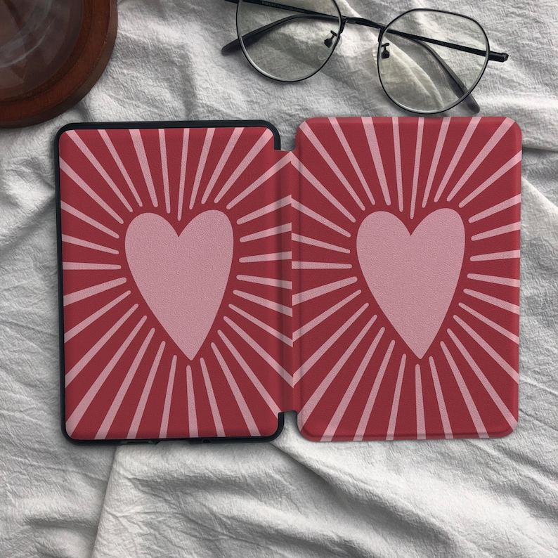 Puede incluir: Una funda rectangular para lector electr&oacute;nico con fondo rojo oscuro y un coraz&oacute;n rosa claro centrado en cada lado. Cada coraz&oacute;n est&aacute; rodeado por l&iacute;neas radiantes de color rosa claro que se extienden hacia los bordes, creando un patr&oacute;n sim&eacute;trico.