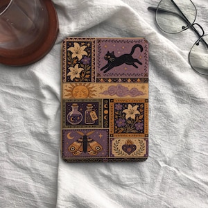 May include: A rectangular e-reader cover featuring a multi-panel illustration in shades of purple, gold, and black. The design includes a leaping black cat, lilies, a sun, clouds, potion bottles, a moth, and a heart with flames in a grid layout.