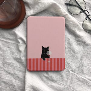 Peut inclure: Une housse de liseuse rectangulaire avec un fond rose clair représentant un chat noir assis à une table à rayures verticales rouges et roses. Le chat est placé à côté d'un verre à vin transparent rempli de vin rouge, centré sur la couverture.