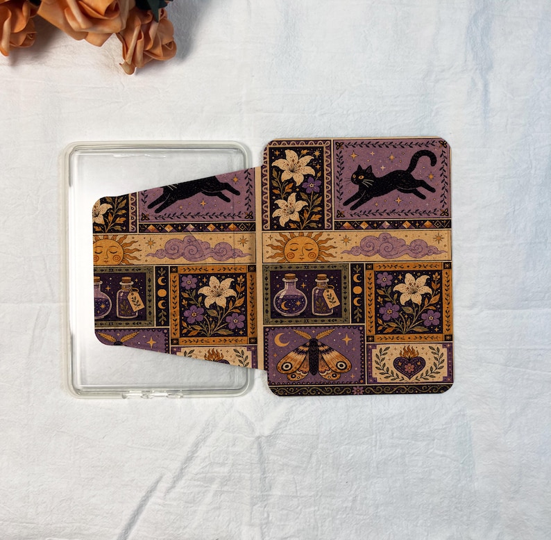 May include: A rectangular clear plastic case featuring a decorative cover with a repeating grid pattern. The design includes illustrations of a black cat, lilies, potion bottles, a sun, clouds, a moth, and a flaming heart in purple and gold.