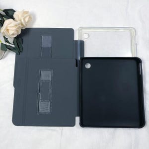 Puede incluir: Una funda para tableta de color gris oscuro con una tapa plegable que presenta una correa el&aacute;stica rectangular y una carcasa interior negra. La funda incluye una secci&oacute;n de marco de pl&aacute;stico transparente con un recorte circular para la c&aacute;mara.