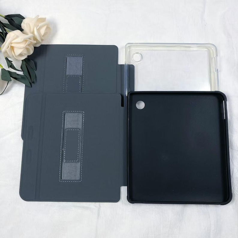 Puede incluir: Una funda para tableta de color gris oscuro con una tapa plegable que presenta una correa el&aacute;stica rectangular y una carcasa interior negra. La funda incluye una secci&oacute;n de marco de pl&aacute;stico transparente con un recorte circular para la c&aacute;mara.