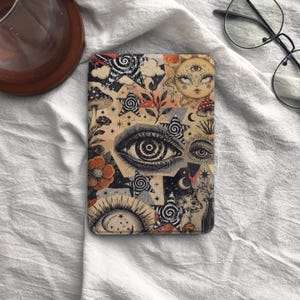 Könnte beinhalten: Eine rechteckige E-Reader-Hülle mit einer Collage aus schwarz-weißen Illustrationen, darunter ein großes zentrales Auge, himmlische Sonnen, Sterne mit Spiralmustern, Pilze und orangefarbene florale Akzente auf einem hellbeigen Hintergrund.