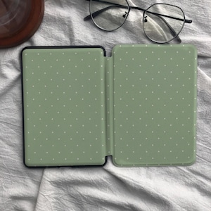Puede incluir: Una funda para lector electr&oacute;nico de color verde salvia con un patr&oacute;n de puntos blancos, mostrada en posici&oacute;n abierta. La funda tiene un marco negro y se exhibe junto a unas gafas de montura fina negra con lentes redondas sobre la cubierta.