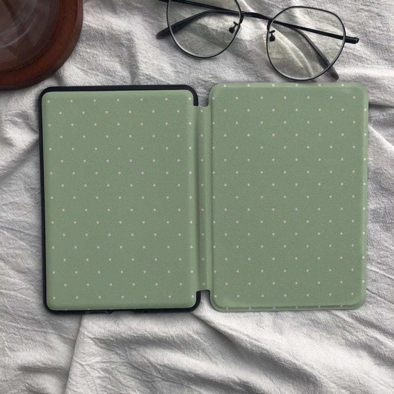 Puede incluir: Una funda para lector electr&oacute;nico de color verde salvia con un patr&oacute;n de puntos blancos, mostrada en posici&oacute;n abierta. La funda tiene un marco negro y se exhibe junto a unas gafas de montura fina negra con lentes redondas sobre la cubierta.
