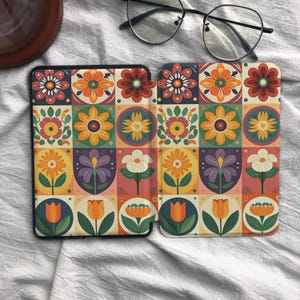 May include: A protective e-reader cover featuring a repeating grid pattern of colorful floral illustrations in shades of orange, yellow, red, purple, and green, with a pair of black-rimmed eyeglasses resting on the fabric above the device.