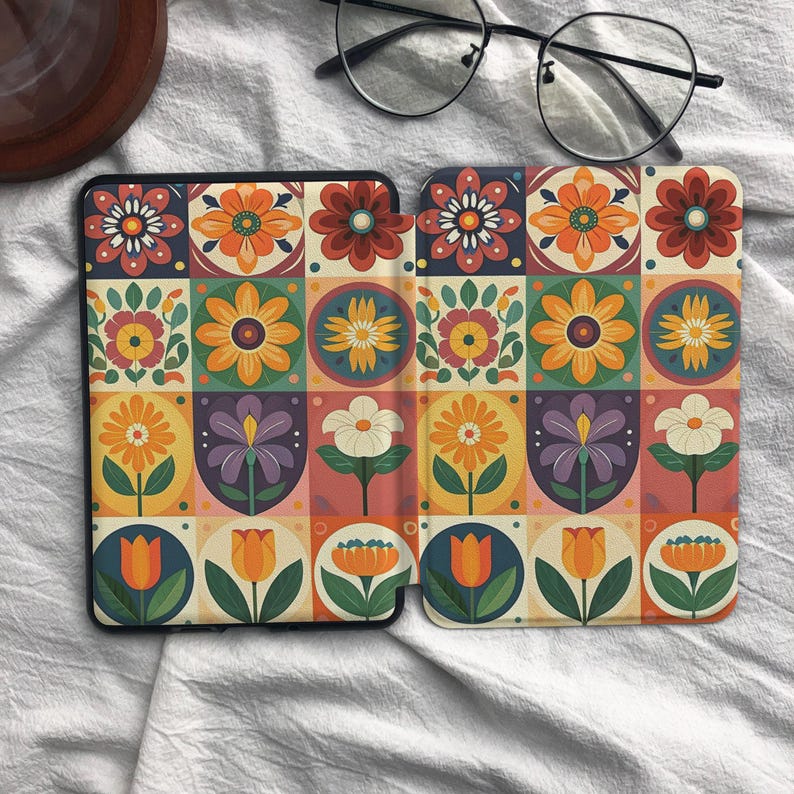May include: A protective e-reader cover featuring a repeating grid pattern of colorful floral illustrations in shades of orange, yellow, red, purple, and green, with a pair of black-rimmed eyeglasses resting on the fabric above the device.