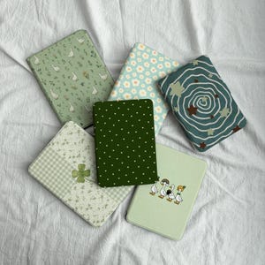 Puede incluir: Seis fundas rectangulares para lector electrónico con varios diseños: un patrón de gansos verde claro, margaritas azul claro, un espiral azul verdoso con estrellas, verde oscuro con corazones blancos, cuadros vichy verde claro y gansos.