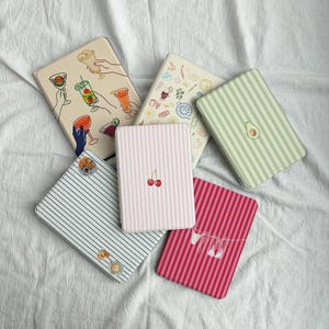 May include: A collection of rectangular cases with various designs. One case features hands holding cocktails, another has a striped pattern with cherries, and others have stripes, an avocado, and food illustrations. The cases are in shades of pink, green, and blue.