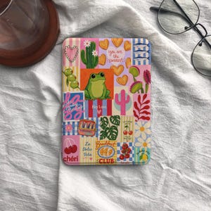 May include: A colorful, patterned tablet case featuring a frog, hearts, cacti, and the text "You are the Sweetest." Other text includes "Sunday Morning," "Hey Love," "chéri," and "La Dolce Vita." The case has a patchwork design with various illustrations.