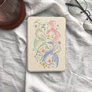 May include: A rectangular e-reader case with a cream-colored background featuring a central illustration of three dragons in pastel green, pink, and purple. The dragons are intertwined with delicate green vines, small colorful flowers, and tiny stars.