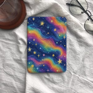 May include: A rectangular e-reader case featuring a vibrant galaxy print with swirling bands of blue, purple, pink, and yellow colors. The surface is decorated with numerous scattered yellow and white stars of varying sizes and shapes.