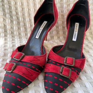 Puede incluir: Un par de zapatos de tac&oacute;n alto rojos y negros con cierres de hebilla. Los zapatos tienen una punta afilada y un tac&oacute;n de aguja. La marca "MANOLO BLAHNIK" est&aacute; impresa en la plantilla.