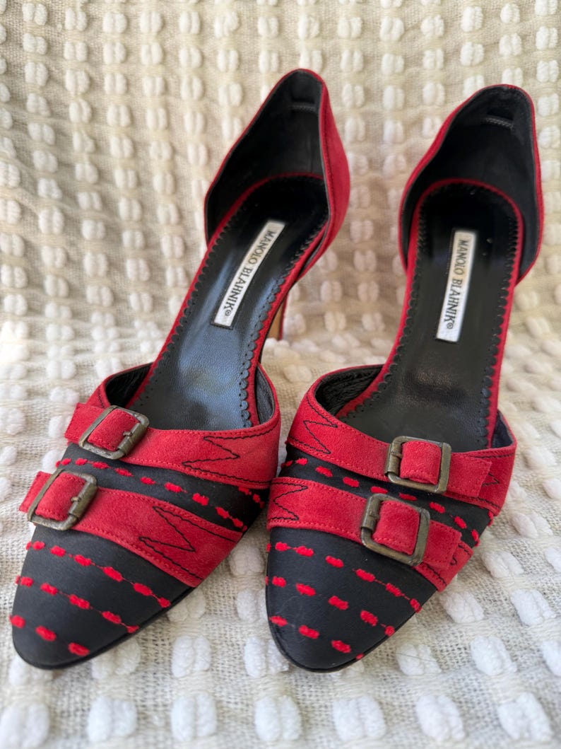 Puede incluir: Un par de zapatos de tac&oacute;n alto rojos y negros con cierres de hebilla. Los zapatos tienen una punta afilada y un tac&oacute;n de aguja. La marca "MANOLO BLAHNIK" est&aacute; impresa en la plantilla.