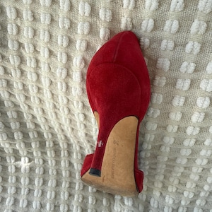 Puede incluir: Zapato de tac&oacute;n alto de ante rojo, con punta afilada y tac&oacute;n de aguja. El zapato tiene la talla 39. El zapato est&aacute; sobre un fondo texturizado blanquecino.