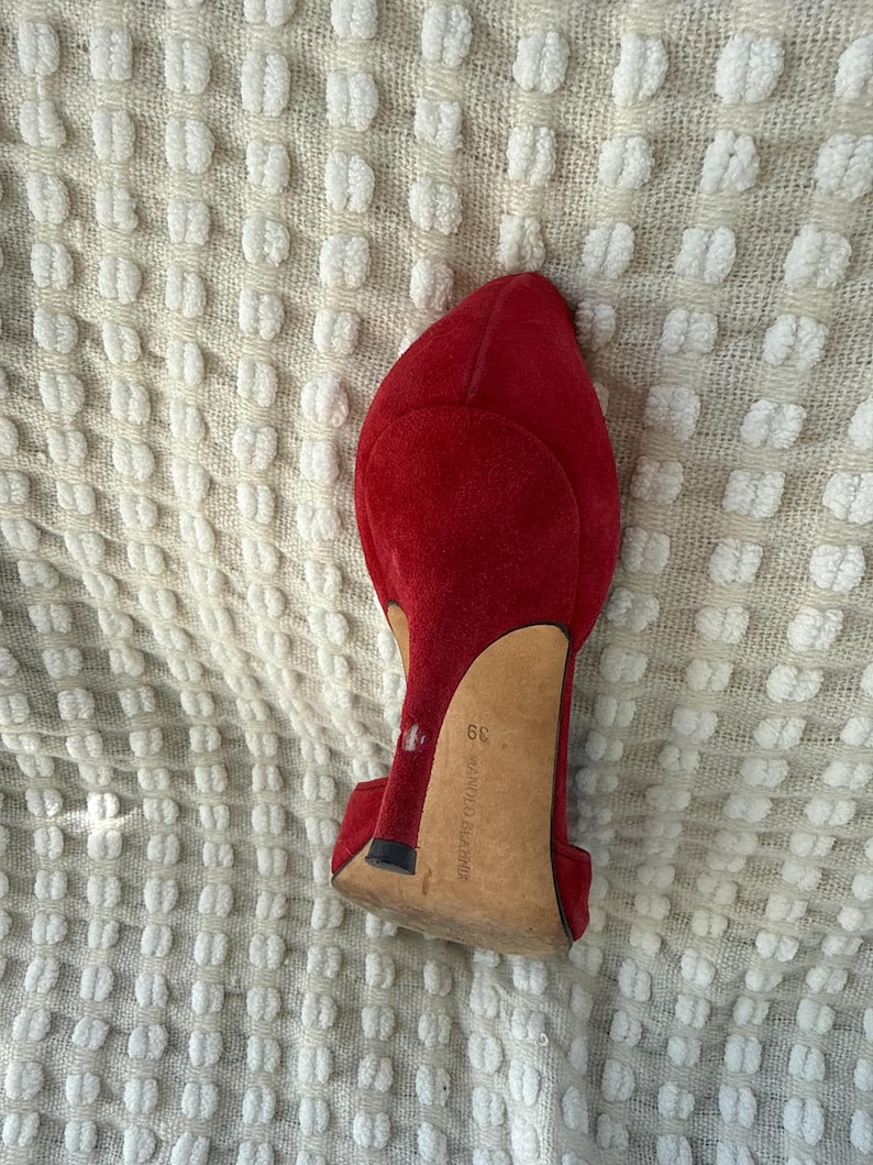 Puede incluir: Zapato de tac&oacute;n alto de ante rojo, con punta afilada y tac&oacute;n de aguja. El zapato tiene la talla 39. El zapato est&aacute; sobre un fondo texturizado blanquecino.