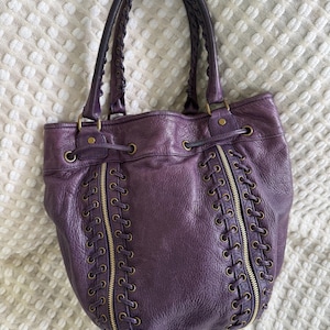 May include: A deep purple leather tote bag with braided handles and gold-toned hardware. The bag features decorative zippers and lace-up detailing on the front. The bag is sitting on a white textured surface.