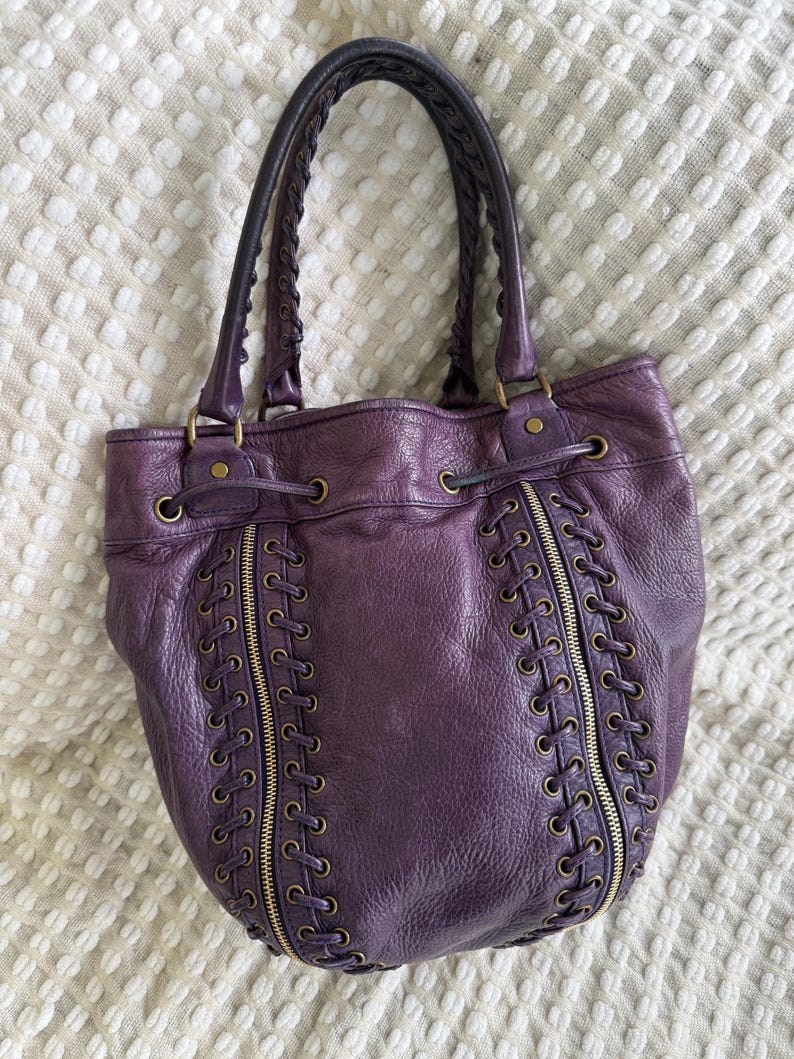May include: A deep purple leather tote bag with braided handles and gold-toned hardware. The bag features decorative zippers and lace-up detailing on the front. The bag is sitting on a white textured surface.
