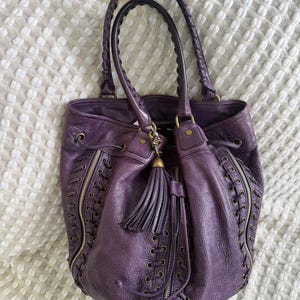 May include: A deep purple leather tote bag with braided handles and a tassel. The bag features decorative zippers and lace-up detailing on the sides. The bag is sitting on a white textured surface.