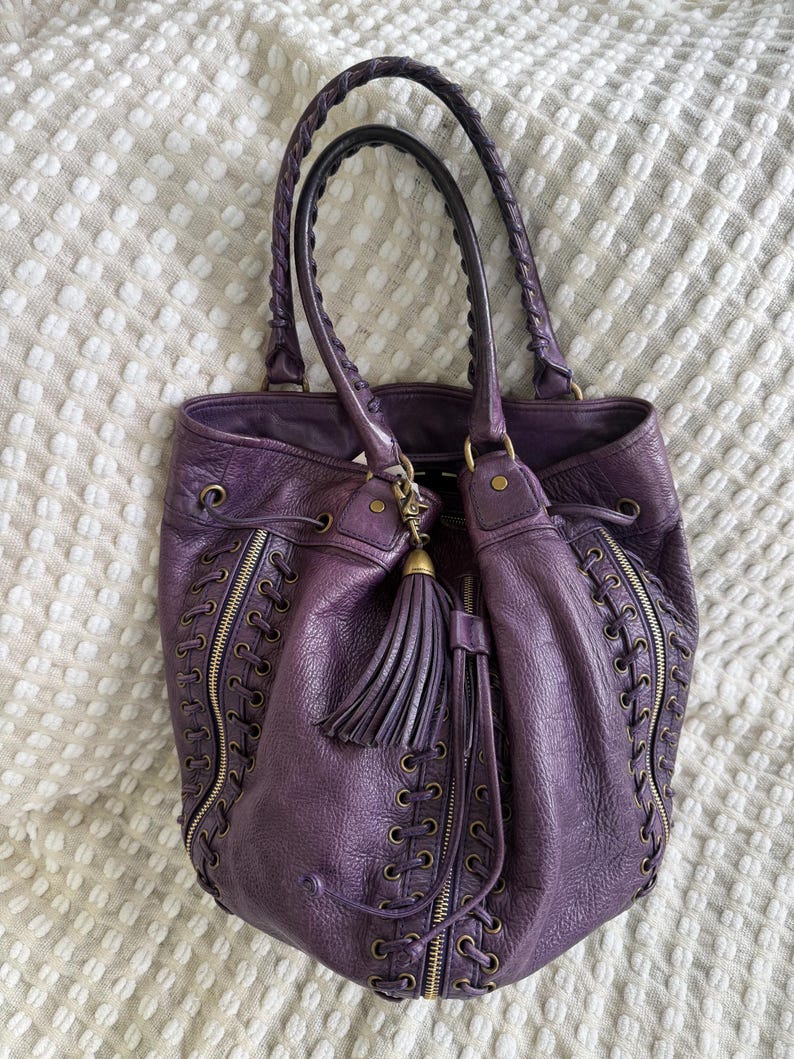 May include: A deep purple leather tote bag with braided handles and a tassel. The bag features decorative zippers and lace-up detailing on the sides. The bag is sitting on a white textured surface.