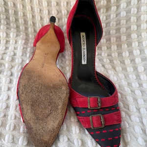 Puede incluir: Un par de tacones Manolo Blahnik de ante rojo con punta y dos correas con hebilla. Los tacones tienen un dise&ntilde;o negro y rojo en la puntera y un tac&oacute;n de aguja. Se ve la suela de un zapato.