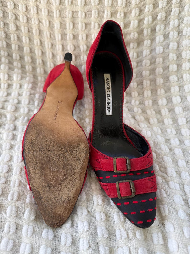 Puede incluir: Un par de tacones Manolo Blahnik de ante rojo con punta y dos correas con hebilla. Los tacones tienen un dise&ntilde;o negro y rojo en la puntera y un tac&oacute;n de aguja. Se ve la suela de un zapato.
