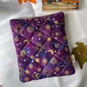 May include: A rectangular quilted fabric pouch featuring a deep purple celestial print with gold suns, moons, and stars. The pouch has a diamond-stitched pattern and a small white fabric tag on the side with the text Queen & Needles printed on it.