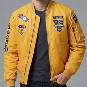 Puede incluir: Chaqueta bomber de color amarillo mostaza con múltiples parches bordados, incluyendo una bomba, un avión y el texto "QUESTION". La chaqueta tiene cierre de cremallera, puños acanalados y el texto "SEALTEAM" en la manga izquierda.
