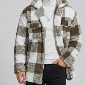 Puede incluir: Una chaqueta de forro polar a cuadros marrones y blancos con cuello y dos bolsillos delanteros. La chaqueta se lleva sobre un cuello alto blanco y pantalones negros. El patrón de la chaqueta es un cuadro grande y audaz.