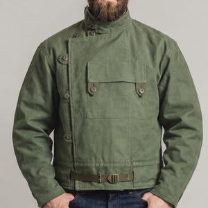 Op de afbeelding: Olijfgroene jas met een hoge kraag, meerdere knopen en een voorzak. De jas heeft een riem in de taille en wordt gecombineerd met een blauwe jeans. Een casual bovenkledingstuk.