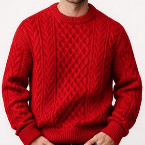 Puede incluir: Un jersey rojo brillante de punto trenzado con cuello redondo. El jersey presenta un patrón de diamantes texturizado en la parte delantera y detalles de trenzas en las mangas. El modelo lleva vaqueros azules.