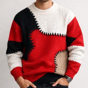May include: A color-block sweater featuring a patchwork design in red, black, white, and tan. The sweater has a crew neck and long sleeves. The knitwear is a mix of cable knit and smooth textures. The model is wearing black jeans and a watch.