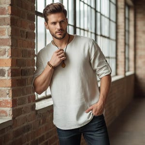 May include: A light gray linen shirt with rolled-up sleeves and a crew neck. The shirt is paired with dark blue jeans. A wooden beaded bracelet and a pendant necklace are also visible.
