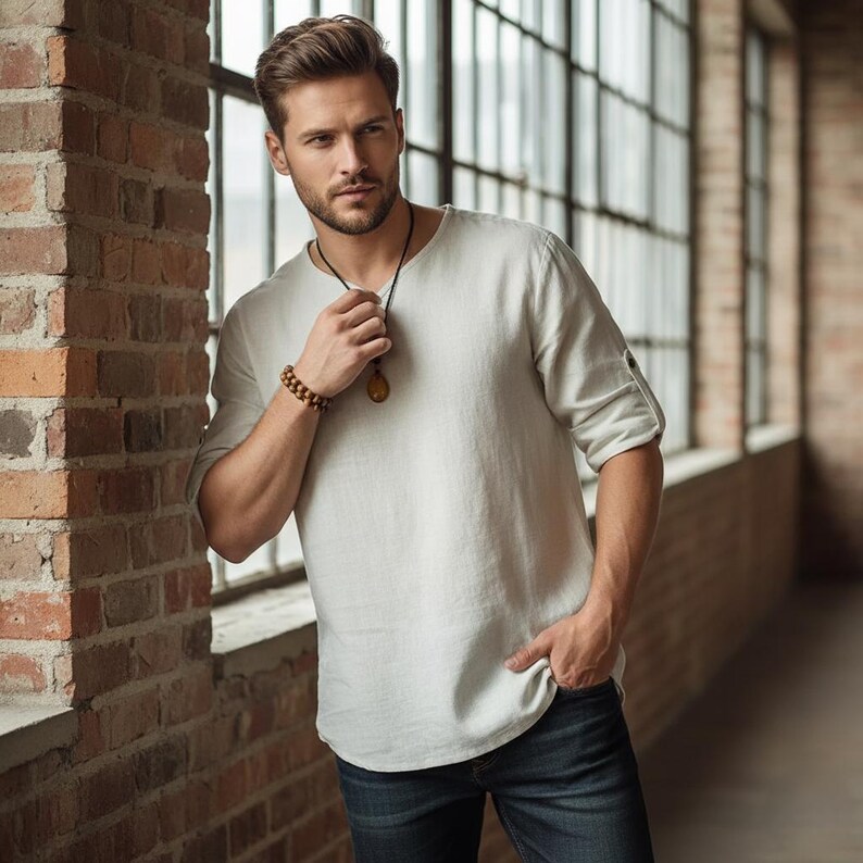 May include: A light gray linen shirt with rolled-up sleeves and a crew neck. The shirt is paired with dark blue jeans. A wooden beaded bracelet and a pendant necklace are also visible.