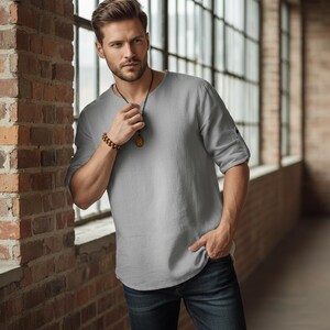 May include: A light gray linen shirt with a v-neck and rolled-up sleeves. The shirt is paired with dark blue jeans. A wooden beaded bracelet and a pendant necklace complete the look. The model is standing near a brick wall and a large window.
